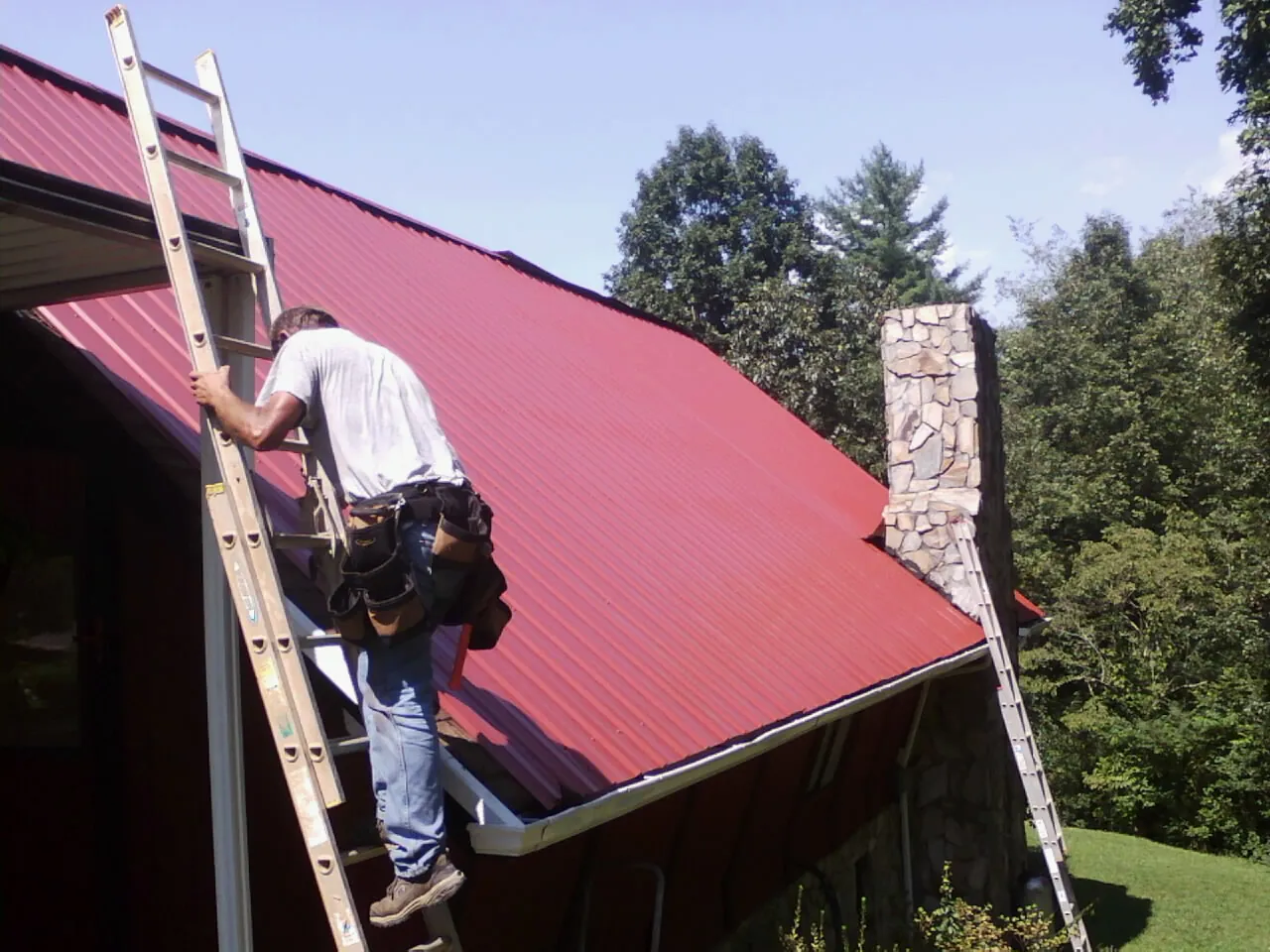 Roofer on a ladder beside a red metal roof and stone chimney — Soffit & Fascia Repair by Roofing Brothers in Tillson